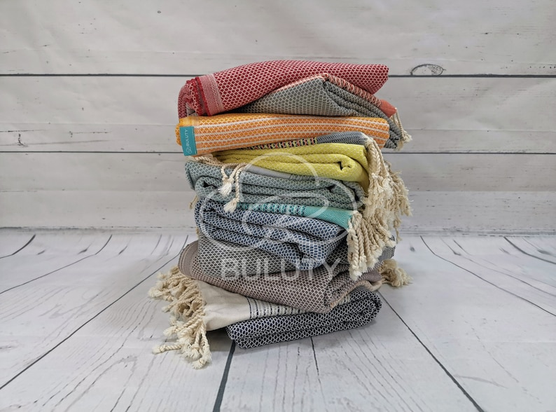 May include: A stack of colorful Turkish towels with fringe, folded and arranged in a pyramid shape. The towels are in shades of red, yellow, green, blue, and gray.