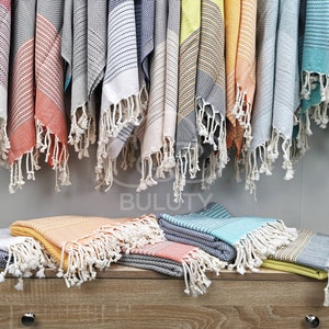 May include: A variety of colorful Turkish towels with fringe, hanging on a wall and folded on a wooden surface. The towels are made of cotton and have a variety of patterns and colors, including blue, green, yellow, orange, and red.