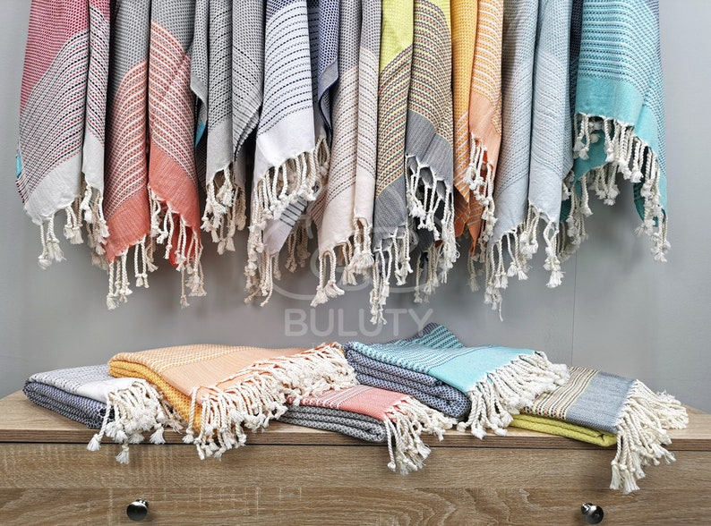 May include: A variety of colorful Turkish towels with fringe, hanging on a wall and folded on a wooden surface. The towels are made of cotton and have a variety of patterns and colors, including blue, green, yellow, orange, and red.