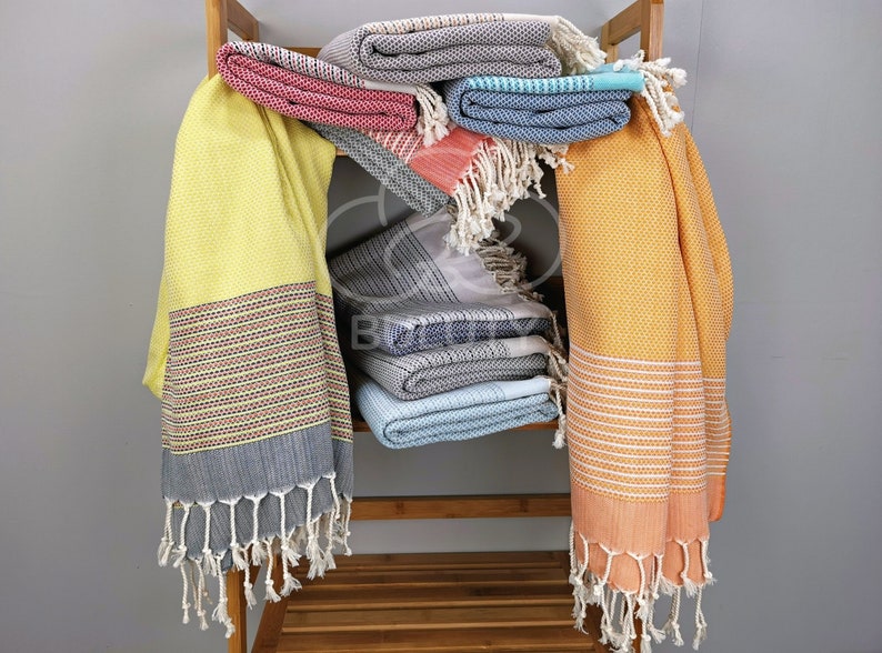May include: A stack of colorful Turkish towels, or peshtemal, with fringe, on a wooden shelf. The towels are in shades of yellow, orange, blue, gray, and pink.