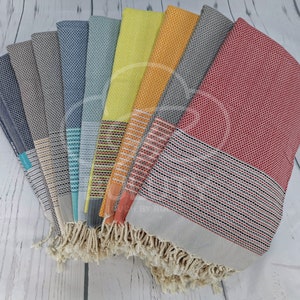 May include: A collection of ten Turkish towels in various colors, including red, orange, yellow, green, blue, and gray. The towels have a woven pattern and fringe on the ends.