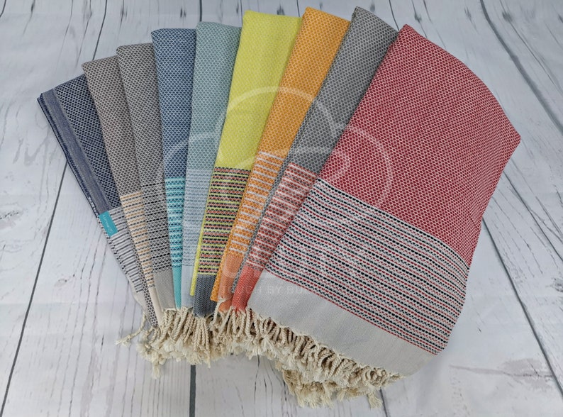 May include: A collection of ten Turkish towels in various colors, including red, orange, yellow, green, blue, and gray. The towels have a woven pattern and fringe on the ends.