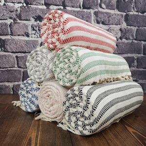 May include: Five rolled up towels with different coloured stripes. The towels are stacked on top of each other on a wooden surface. The towels are made of cotton and have a soft texture.