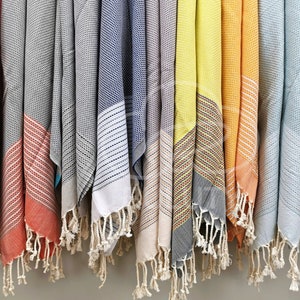 May include: A row of colorful Turkish towels with fringe hanging from a rack. The towels are in various shades of red, gray, blue, yellow, orange, and teal.