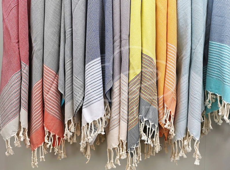 May include: A row of colorful Turkish towels with fringe hanging from a rack. The towels are in various shades of red, gray, blue, yellow, orange, and teal.