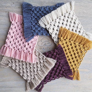 May include: Five handmade crocheted coasters with fringe in various colors, including pink, beige, blue, white, and yellow. The coasters are arranged in a circle on a wooden surface.