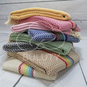 May include: A stack of six colorful patterned towels with fringe edges. The towels are folded and stacked on a white wooden surface. The towels are in various colors, including yellow, pink, green, blue, and brown.