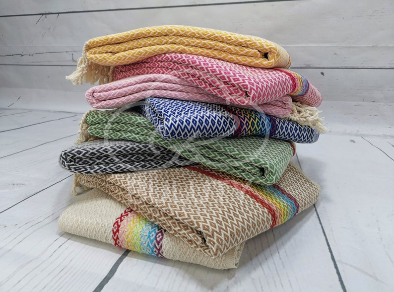 May include: A stack of six colorful patterned towels with fringe edges. The towels are folded and stacked on a white wooden surface. The towels are in various colors, including yellow, pink, green, blue, and brown.
