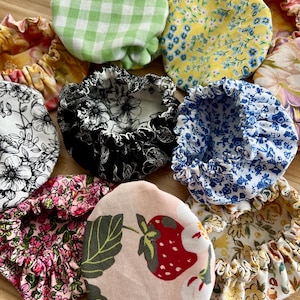 Pode incluir: Uma coleção de capas de tecido para tigelas em vários padrões. Os desenhos incluem xadrez, floral e uma estampa de morango. As capas são em tons de verde, amarelo, azul, rosa, preto e branco. As bordas são franzidas.