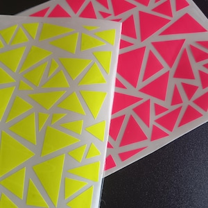 Op de afbeelding: Twee vellen stickers met een geometrisch ontwerp. Eén vel heeft neongele driehoeken en de andere heeft neonroze driehoeken. De driehoeken zijn in een willekeurig patroon op een transparante achtergrond gerangschikt.