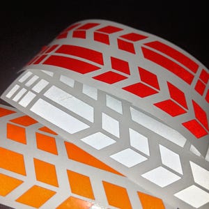 Op de afbeelding: Drie rollen reflecterende tape met een geometrisch patroon. De tape is oranje, wit en rood.