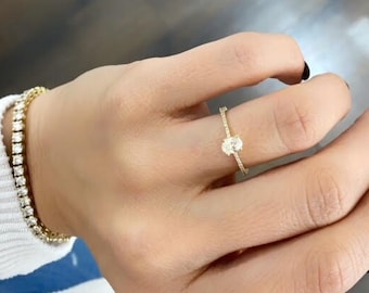 May include: A gold ring with a single oval-shaped diamond set in the center. The ring is surrounded by smaller diamonds. The ring is on a person's finger.