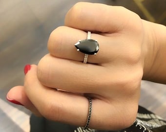 May include: A pear-shaped black diamond engagement ring with a pave diamond band. The ring is on a finger with red nail polish.