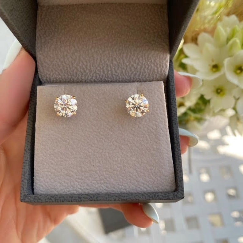 May include: A pair of round-cut diamond stud earrings in a gray jewelry box. The earrings are set in gold and sparkle brightly. The box is held in a hand, with flowers in the background.