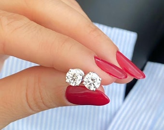 May include: A pair of round diamond stud earrings in a white gold setting. The earrings are shown on a person's finger with red nail polish.