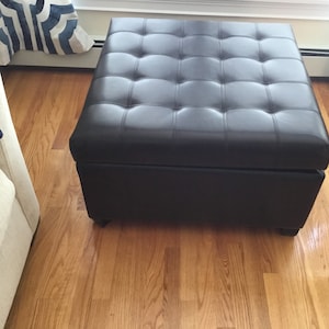 May include: A brown leather storage ottoman with a tufted top. The ottoman is sitting on a hardwood floor.