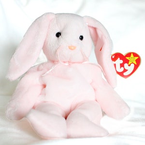 May include: Pink plush bunny with long floppy ears and a white ribbon around its neck. The bunny has a red and white Ty tag attached to its ear that reads "Beanie Babies Baby".