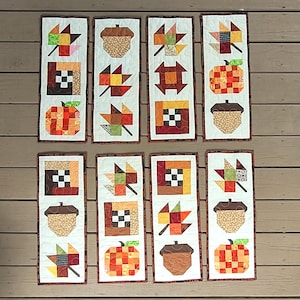 May include: Eight rectangular quilted table runners featuring autumn-themed patchwork designs. Each runner displays a vertical arrangement of fabric blocks depicting colorful maple leaves, acorns, pumpkins, and checkered squares in shades of orange, brown, yellow, and green.