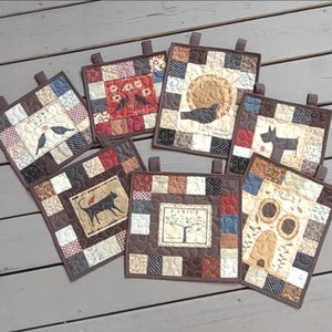 May include: Six square quilted wall hangings featuring patchwork borders in shades of brown, cream, and red. Each piece displays a central graphic including birds, a dog, a cat, a tree labeled FAMILY, and a beehive with the text TODAY.