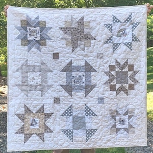 May include: A square quilt featuring a nine-block grid pattern in shades of cream, gray, and brown. The quilt displays various star and geometric block designs, some incorporating patterned fabrics with small illustrations, all finished with a cream border.