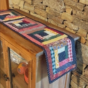 May include: A rectangular table runner featuring a patchwork log cabin design with various fabric patterns including red and white gingham, floral prints, stripes, and solid colors in shades of blue, yellow, green, pink, and brown with a dark blue border.