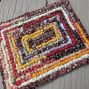 May include: A handmade, square braided rug with a patchwork design. The rug features a variety of fabrics in shades of brown, red, yellow, and white. The braided strips are arranged in concentric squares, creating a textured surface. The rug is likely used as a decorative accent or a small floor covering.