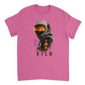 Könnte beinhalten: Ein rosafarbenes Kurzarm-T-Shirt mit einer Grafik eines Astronautenhelms, in dessen Visier eine Sonnenuntergangslandschaft zu sehen ist. Unter dem Helm steht der Text: SOME SECRETS SHOULD STAY BURIED. SILO ist in großen schwarzen Buchstaben gedruckt.