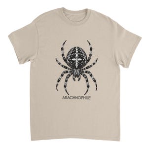 Op de afbeelding: Een beige t-shirt met korte mouwen voorzien van een gedetailleerde zwart-witillustratie van een spin met een kruisvormig patroon op het achterlijf. Onder de spin staat het woord ARACHNOPHILE in een eenvoudig, donkergrijs schreefloos lettertype.