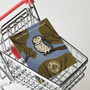 Könnte beinhalten: Ein silberner Einkaufswagen mit rosa Griff hält eine Karte mit einem Eulen-Anstecker. Der Anstecker ist gold-schwarz, und die Karte trägt den Text "SMALL PIN - BIG STATEMENT". Der Eulen-Anstecker befindet sich auf einem blau-braunen Hintergrund.