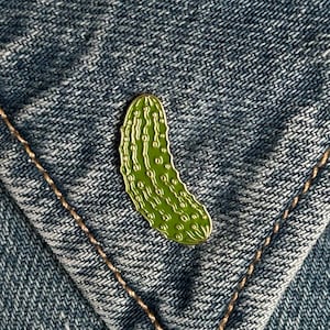 Może przedstawiać: Zielona przypinka w kształcie ogórka z białymi detalami, na niebieskim tle z dżinsu. Przypinka jest przymocowana do kieszeni kurtki dżinsowej. Przypinka to zabawny dodatek.