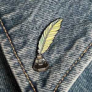 May include: A feather and inkwell enamel pin with yellow, green, and black details. The pin is attached to a blue denim jacket. The feather has a gradient of yellow to green. The inkwell is black.