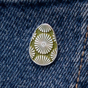 May include: An egg-shaped enamel pin with a white radial design on a green background. The pin is set against a blue denim background, showcasing the intricate details of the design. The pin is a decorative accessory.