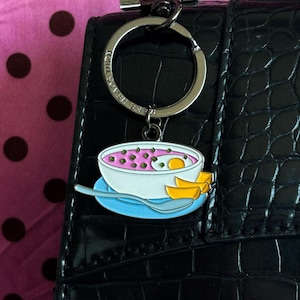 May include: A keychain with a food-themed charm. The charm features a bowl of soup with a pink broth, green dots, and a yellow egg. The charm is attached to a silver keyring and a black leather strap.