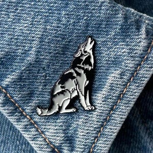 May include: A silver and black enamel pin in the shape of a howling wolf. The pin is attached to a denim jacket. The wolf is depicted in a seated position with its head raised and mouth open, as if howling.