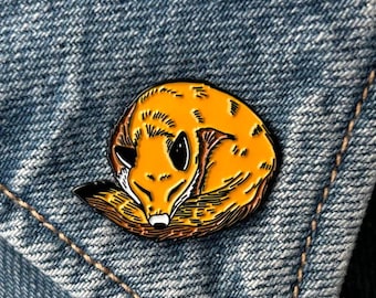 May include: A round enamel pin featuring a curled-up fox in shades of orange, brown, and black. The fox pin is set against a blue denim background, with the pin attached to the denim fabric.