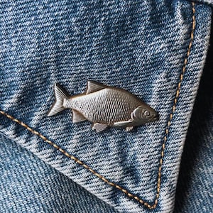 May include: A silver-colored fish-shaped pin is affixed to the denim fabric of a jacket. The pin is detailed with scales and fins. The jacket is blue denim with gold stitching.