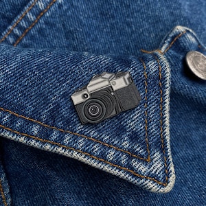 Puede incluir: Un pin de esmalte de cámara negro y plateado en una chaqueta vaquera azul.