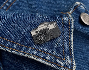 Puede incluir: Un pin de esmalte de cámara negro y plateado en una chaqueta vaquera azul.