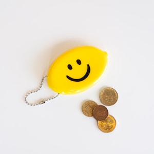 May include: A yellow coin purse with a smiling face design and a silver chain. Several coins are scattered nearby. The purse is oval-shaped and has a simple, cheerful design.