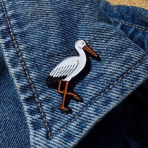 Könnte beinhalten: Ein weiß-schwarzer Emaille-Storchen-Anstecker mit orangefarbenen Beinen und Schnabel, auf einer blauen Jeansjacke. Der Anstecker zeigt ein detailliertes Design des Vogels in einer Laufpose mit glänzendem Finish.
