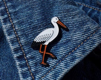 May include: A white and black enamel stork pin with orange legs and beak, set against a blue denim jacket. The pin features a detailed design of the bird in a walking pose, with a glossy finish.