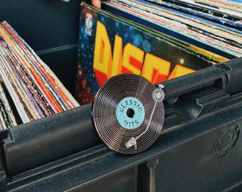 Puede incluir: Una caja de almacenamiento de plástico negro llena de discos de vinilo. Un pin de esmalte plateado y azul con forma de tocadiscos está en el borde de la caja. El pin dice "Classic Hits" en el disco.