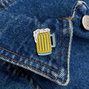 May include: A yellow beer mug enamel pin with white foam, a blue base, and a blue handle. The pin is set against a blue denim jacket.