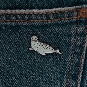 May include: A silver-toned metal enamel pin shaped like a spotted seal, featuring a light gray body with dark gray spots, whiskers, and a smooth finish, attached to a piece of dark blue denim fabric with visible orange stitching along the seam.