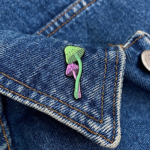 Może przedstawiać: Szpilka z emaliowanego grzyba w kolorze zielonym i różowym na niebieskiej dżinsowej kurtce.