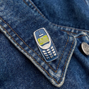 May include: A blue and silver enamel pin shaped like a vintage cell phone with a green screen and a keypad. The pin is on a blue denim jacket.