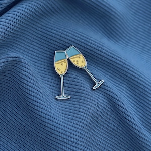 Könnte beinhalten: Eine silberne Emaille-Anstecknadel mit zwei Champagnergläsern, die anstoßen. Die Gläser sind mit gelber Flüssigkeit gefüllt und haben blaue Ränder.
