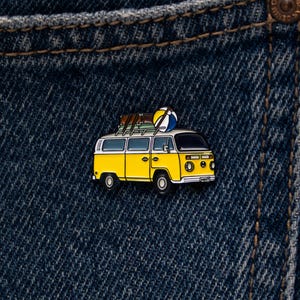 May include: A yellow and white enamel pin shaped like a vintage camper van. The van features a roof rack carrying a brown suitcase and a blue, yellow, and white beach ball. The van has black wheels, dark windows, and a small white license plate.