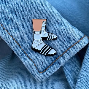 May include: An enamel pin featuring a cartoon leg wearing white socks and black sandals.