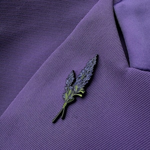 May include: A lavender-themed lapel pin with blue and green accents, resting on a purple blazer. The pin features a detailed design of lavender sprigs, adding a touch of nature-inspired elegance to the garment. The blazer's fabric has a subtle texture.